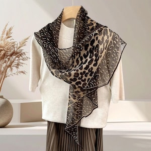 May include: A sheer scarf with a leopard print design in black and tan. The scarf is draped over a cream-colored sweater and a pleated skirt. The scarf has a dark border and a repeating pattern of curved shapes.
