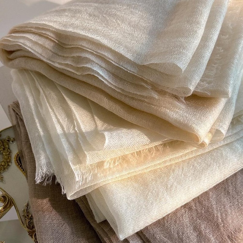 May include: A stack of folded scarves in shades of cream and light beige. The fabric has a fine, semi-sheer texture with visible woven threads and delicate, frayed edges along the borders, creating a soft, layered appearance in the light.