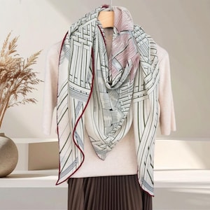 May include: A cream scarf featuring a geometric design in black, green, and red. The scarf is draped over a light-coloured sweater on a wooden hanger. The scarf has a red border. The image showcases a fashion item.