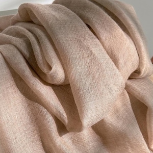 May include: A lightweight, semi-sheer scarf in a soft, muted beige or light tan hue. The fabric features a fine, woven texture with visible threads and loose, flowing folds that create depth and shadows across the surface of the material.