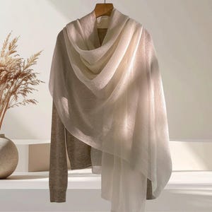 May include: A long-sleeved beige sweater layered with a sheer, off-white scarf draped over the shoulders. The sweater features a ribbed knit texture, while the scarf is made of a lightweight, semi-transparent fabric with soft, flowing folds.