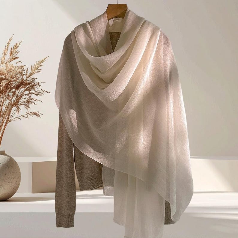 May include: A long-sleeved beige sweater layered with a sheer, off-white scarf draped over the shoulders. The sweater features a ribbed knit texture, while the scarf is made of a lightweight, semi-transparent fabric with soft, flowing folds.