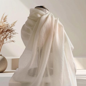 May include: A soft, off-white scarf draped over a display form. The fabric has a lightweight, semi-sheer appearance with a fine, woven texture. The scarf hangs in loose, flowing folds, creating a layered look that highlights the material's drape.