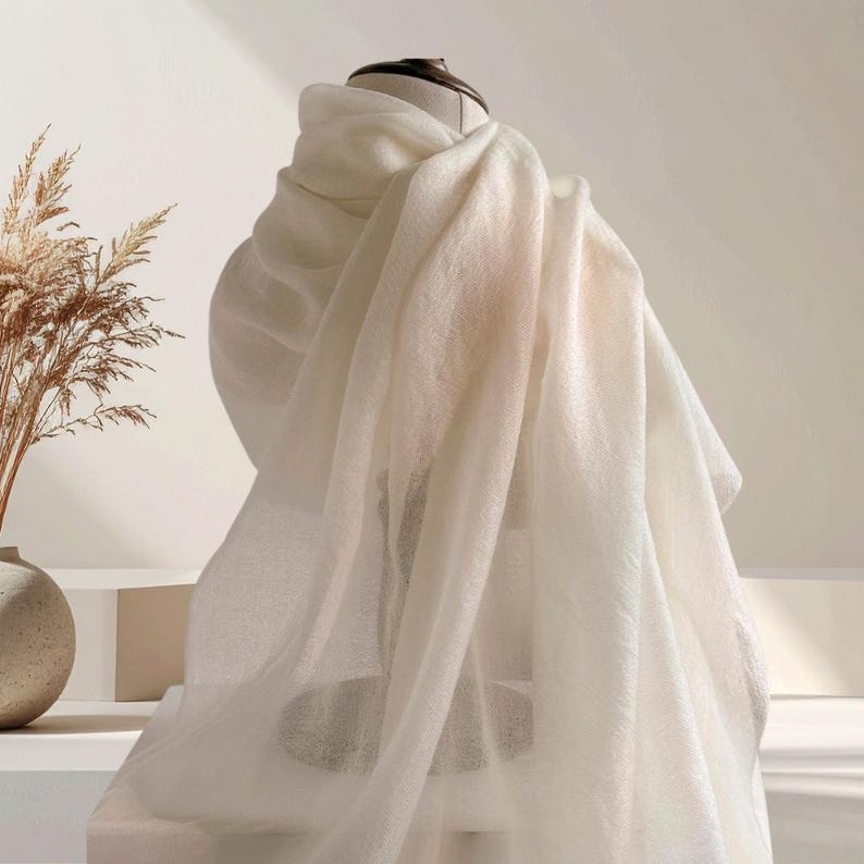 May include: A soft, off-white scarf draped over a display form. The fabric has a lightweight, semi-sheer appearance with a fine, woven texture. The scarf hangs in loose, flowing folds, creating a layered look that highlights the material's drape.