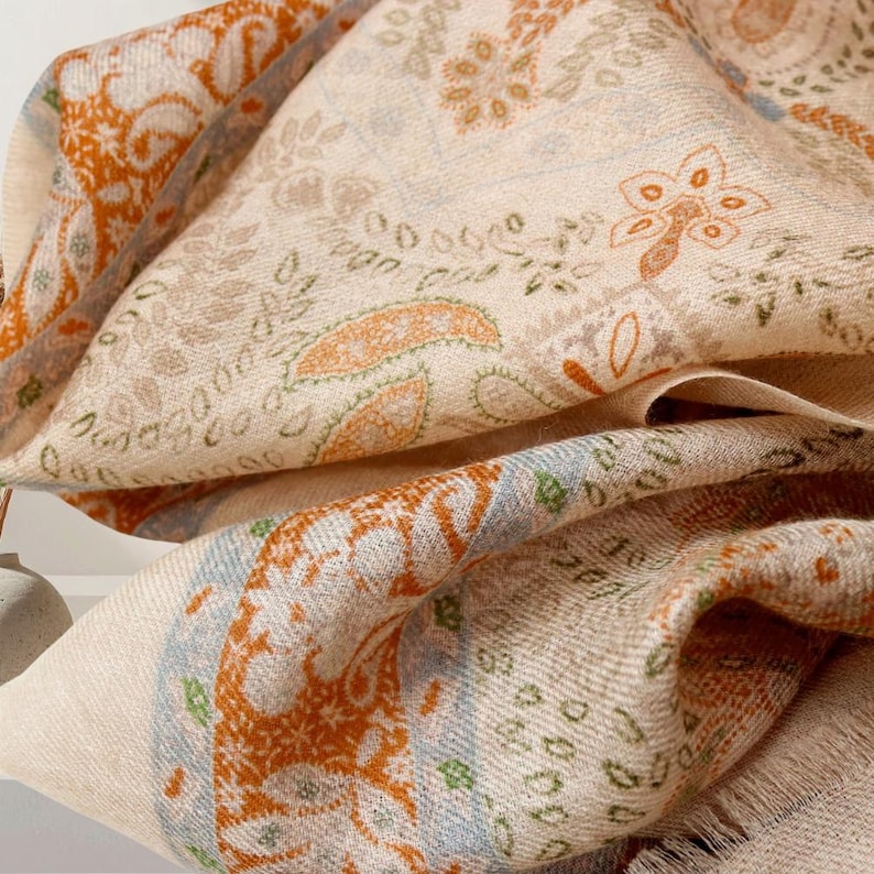 Op de afbeelding: Een beige sjaal met een ingewikkeld paisley-patroon in tinten oranje, groen en blauw. De sjaal is opgevouwen, waardoor het gedetailleerde ontwerp en de gefranjerde randen zichtbaar zijn. De algehele uitstraling is zacht en getextureerd.