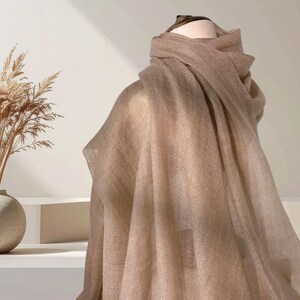 May include: A lightweight, semi-sheer scarf in a solid, neutral beige tone. The fabric features a fine, woven texture and is draped elegantly over a mannequin form, creating soft, cascading folds that highlight the material's delicate quality.