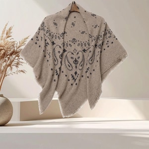 May include: A beige poncho featuring a dark brown paisley and geometric bandana-style print. The garment has a soft, textured appearance with a draped silhouette and frayed edges along the bottom hem, hanging from a wooden hanger.