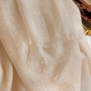 May include: A soft, off-white scarf with a fine, woven texture and delicate frayed edges. The fabric displays a subtle, light-reflecting sheen across its surface, with multiple folds creating depth and highlighting the lightweight, airy quality.