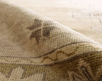 May include: Close-up of a beige rug with a rolled edge. The rug features a detailed pattern in shades of brown and green. The texture of the rug is visible, showcasing the woven fibres and intricate design. The rug's colour palette is neutral, making it suitable for various interior styles.