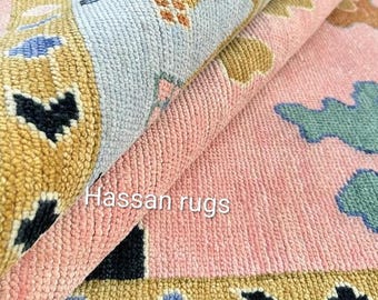 Könnte beinhalten: Nahaufnahme eines aufgerollten Teppichs mit einem rosa- und hellblauen Blumendesign. Der Teppich hat einen goldenen Rand mit schwarzen, blauen und goldenen geometrischen Formen. Der Text "Hassan rugs" ist auf dem Teppich zu sehen.