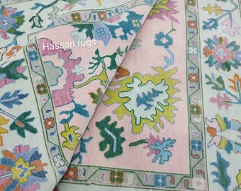 Könnte beinhalten: Ein aufgerollter Teppich mit einem Blumenmuster in Rosa-, Blau-, Grün-, Gelb- und Orangetönen. Der Teppich hat einen cremefarbenen Rand und eine rosa Mitte. Der Text "Hassan rugs" ist auf dem Teppich zu sehen.
