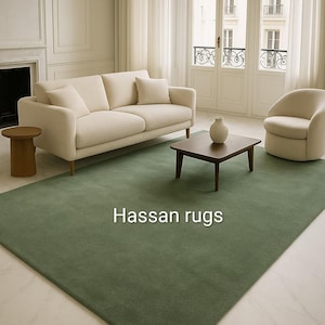 May include: A rectangular sage green rug with the text "Hassan rugs" in white. The rug is in a living room setting with a cream-colored sofa, chair, and coffee table. A white vase sits on the table.