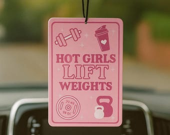 May include: Pink car air freshener with the text "HOT GIRLS LIFT WEIGHTS". The design includes dumbbell, shaker, weight plate, and kettlebell illustrations. The weight plate shows 20 lbs. The air freshener hangs from a black string.