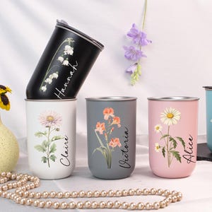 Puede incluir: Una colección de vasos de acero inoxidable personalizados en varios colores, cada uno adornado con diseños florales y nombres. Los vasos incluyen negro, blanco, gris, rosa y azul, con nombres como Hannah, Claire, Victoria, Alice y Harper.