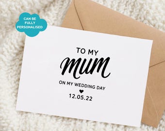 Peut inclure: Une carte de vœux rectangulaire blanche avec le texte noir TO MY mum ON MY WEDDING DAY suivi d'un petit cœur et de la date 12.05.22. Une enveloppe marron clair est glissée derrière la carte, qui présente une bulle bleue avec du texte.