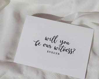 May include: A rectangular white card featuring black printed text that reads "will you be our witness?" in a cursive script, with the name "EVELYN" printed in a simple sans-serif font directly underneath the main question on the card.
