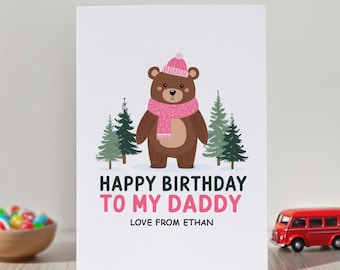 Op de afbeelding: Een witte verjaardagskaart met een bruine beer die een roze gebreide muts en een bijpassende roze sjaal draagt. De beer staat tussen vier groene dennenbomen. De tekst luidt: HAPPY BIRTHDAY TO MY DADDY LOVE FROM ETHAN in het zwart en roze.