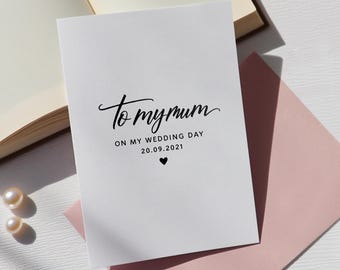 Peut inclure: Une carte de vœux rectangulaire blanche avec un texte noir indiquant To my mum ON MY WEDDING DAY 20.09.2021 et une petite icône de cœur noir. La carte est placée à côté d'une enveloppe rose clair et d'un livre aux pages crème.