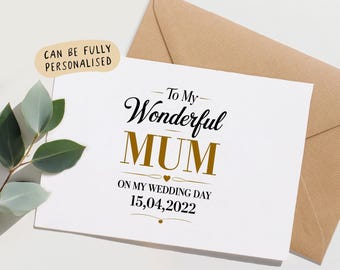 Peut inclure: Une carte de vœux blanche avec du texte noir et doré indiquant To My Wonderful MUM ON MY WEDDING DAY 15,04,2022. Une enveloppe beige est placée derrière la carte et un petit brin de feuilles vertes est disposé à gauche de la carte.