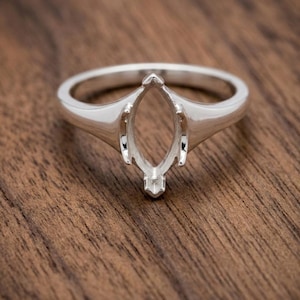 Könnte beinhalten: Eine silberne Ringfassung mit einem marquiseförmigen Mittelteil. Der Ring hat eine polierte Oberfläche und ein klassisches Design, perfekt für einen individuellen Edelstein. Der Ring wird auf einer Holzoberfläche gezeigt.