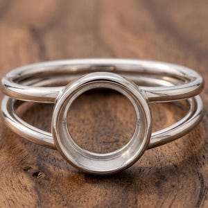 May include: A silver ring with a circular setting, designed to hold a gemstone. The ring features a double-band design, with a polished finish. The ring is set against a wooden background.