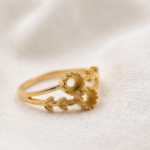May include: A gold-toned ring featuring a split band design. One side of the band displays a delicate leaf motif, while the other side features two circular, gear-like settings intended for small stones or decorative accents.