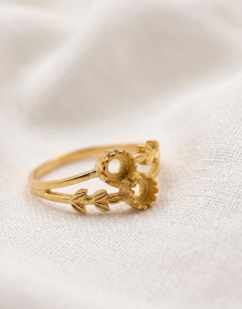 May include: A gold-toned ring featuring a split band design. One side of the band displays a delicate leaf motif, while the other side features two circular, gear-like settings intended for small stones or decorative accents.