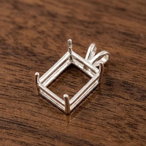 May include: A silver rectangular pendant setting with four prongs and a loop for a chain. The setting is designed to hold a gemstone. The pendant is displayed on a dark wood surface.