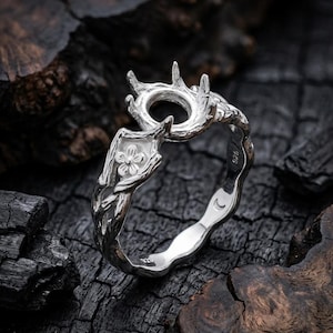 May include: A silver ring with a nature-inspired design. The ring features a floral accent and a prong setting, with a textured band. The ring is set against a dark, textured background.