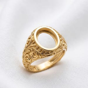 May include: A gold-colored ring with an oval setting and ornate, swirling floral engravings. The ring's band and setting are a consistent gold tone, and the design is symmetrical. The ring is set against a white background.