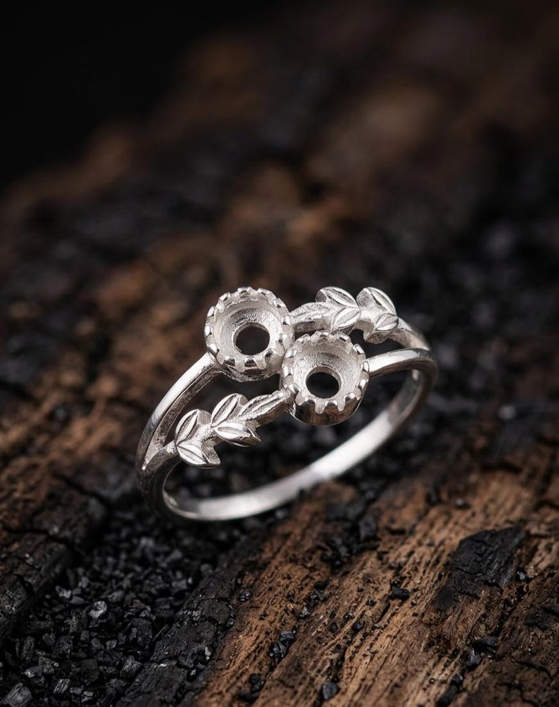 May include: A silver ring featuring two circular prong settings for stones, arranged side by side. The band is decorated with detailed leaf motifs extending from the settings along the sides of the ring, creating a nature-inspired design.