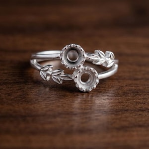 May include: A silver ring featuring two circular prong settings for stones, arranged in a bypass style. The band is decorated with detailed leaf motifs extending from each setting, creating a nature-inspired design in polished metal.