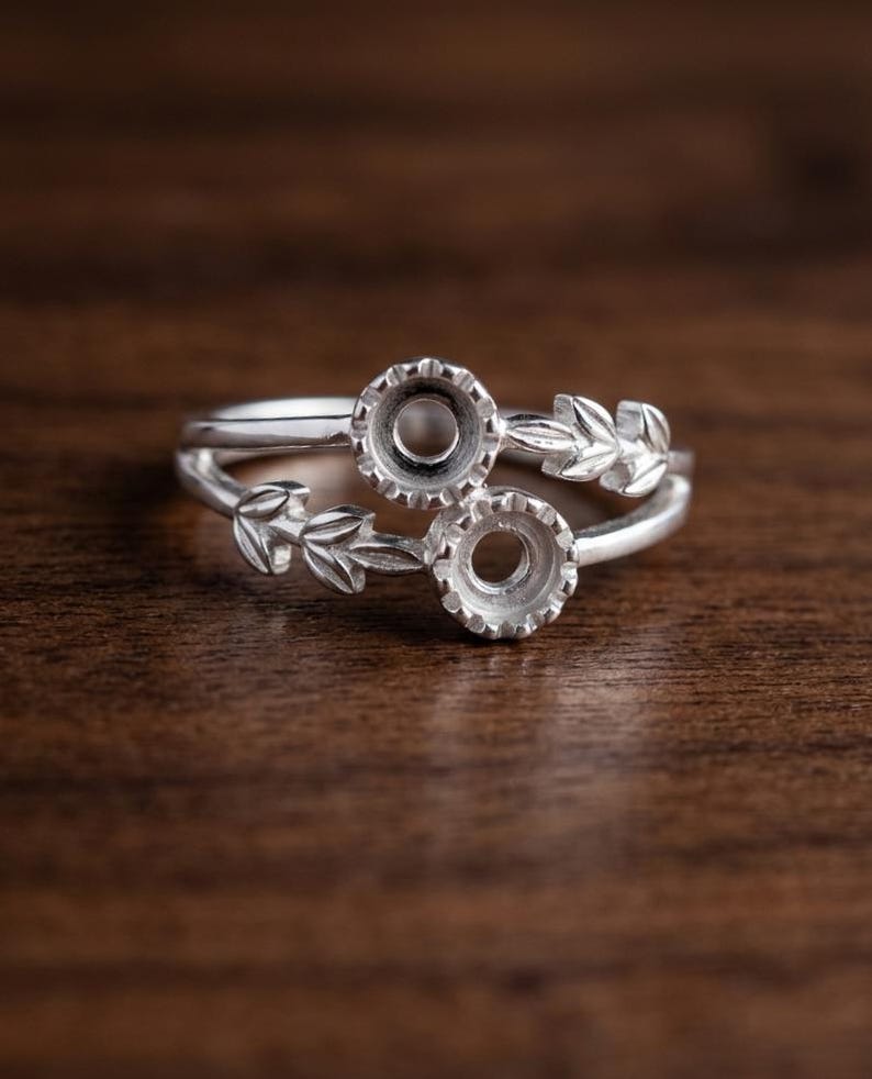 May include: A silver ring featuring two circular prong settings for stones, arranged in a bypass style. The band is decorated with detailed leaf motifs extending from each setting, creating a nature-inspired design in polished metal.