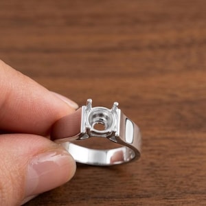 May include: A silver ring setting with a rectangular band and a circular bezel designed to hold a gemstone. The setting has two prongs and a polished finish, ideal for jewelry making.