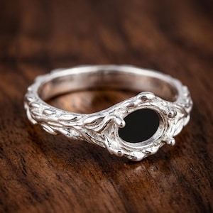 May include: A silver ring with an oval black stone setting. The ring band features a detailed, nature-inspired design with leaf and vine motifs. The ring is displayed on a dark wood surface.