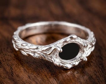 May include: A silver ring with an oval black stone setting. The ring band features a detailed, nature-inspired design with leaf and vine motifs. The ring is displayed on a dark wood surface.