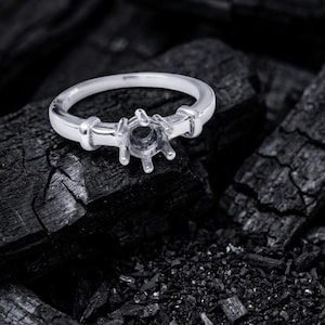 May include: A silver ring with a prong setting, designed to hold a gemstone. The ring is set against a dark, textured background, likely charcoal. The ring's band is smooth and simple, with a detailed setting.