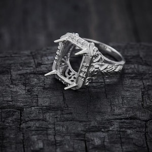 May include: A silver ring setting featuring a rectangular head with four pointed prongs designed to hold a gemstone. The band and the gallery of the ring are decorated with intricate, raised metalwork patterns along the sides and shoulders.