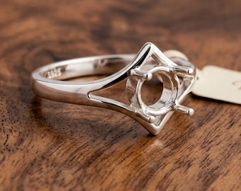 May include: A silver ring setting with a geometric design. The ring features a central round setting and a decorative, openwork design. The ring is made of a shiny, silver-colored metal and is displayed on a wooden surface.