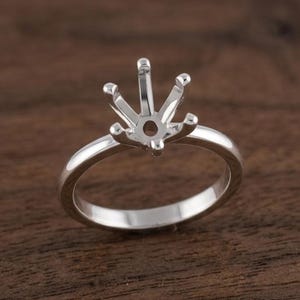 May include: A silver ring setting with a six-prong design for holding a gemstone. The ring features a polished band and a detailed setting, ideal for a custom jewellery piece. The ring is displayed on a dark wood surface.