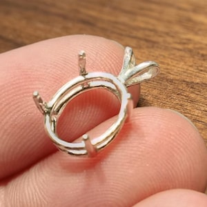 Peut inclure: Un sertissage de bijoux de couleur argentée, de forme ovale, avec quatre griffes et une bélière décorative. Le sertissage est conçu pour maintenir une pierre précieuse. Il est tenu dans une main.