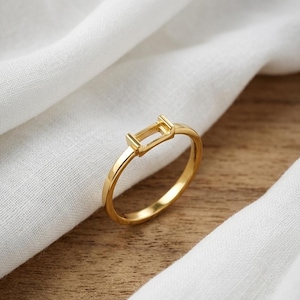 May include: A gold-toned ring featuring a thin, polished band and a rectangular, open-frame setting at the top. The setting is designed to hold a stone or decorative element, showcasing a smooth, metallic finish throughout the entire piece.