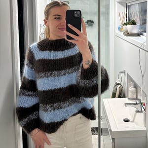 May include: A striped jumper in brown and pale blue, featuring a high neck and voluminous sleeves. The person in the image is wearing the jumper with light-coloured trousers. The jumper's design is a classic, comfortable style.