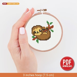 Pode incluir: Um bastidor de bordado em ponto cruz com um bicho-preguiça sorridente pendurado num galho. O bicho-preguiça é castanho com um rosto castanho claro e folhas verdes. O bastidor tem 7,5 cm de diâmetro e tem um rótulo de download PDF.