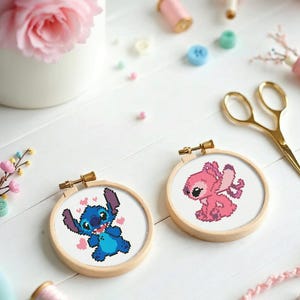 May include: Two cross-stitch hoops featuring the characters Stitch and Angel from Lilo & Stitch. Stitch is blue with hearts, Angel is pink. The hoops are in wooden frames, surrounded by craft supplies like scissors, thread, and flowers.