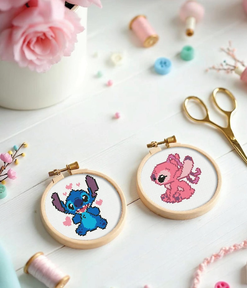 May include: Two cross-stitch hoops featuring the characters Stitch and Angel from Lilo & Stitch. Stitch is blue with hearts, Angel is pink. The hoops are in wooden frames, surrounded by craft supplies like scissors, thread, and flowers.