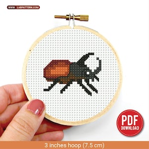 Könnte beinhalten: Ein Kreuzstich-Stickrahmen mit einem pixeligen Käfer-Design. Der Käfer ist braun und schwarz, vor einem weißen Hintergrund. Der Holzrahmen hat einen Durchmesser von 7,5 cm. Das Bild enthält den Text "PDF DOWNLOAD".