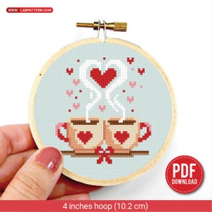 Puede incluir: Un bordado de punto de cruz en un bastidor de madera. El diseño presenta dos tazas de café con vapor en forma de corazón, formando un corazón. Las tazas tienen corazones rojos y el fondo es azul claro. El bastidor tiene 10,2 cm de diámetro.