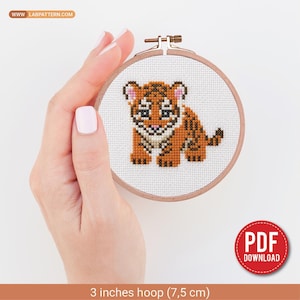 May include: A cross-stitch pattern of a small orange and black tiger cub centered in a 3-inch wooden embroidery hoop. The design features a white background with the text WWW.LABPATTERN.COM at the top and a red PDF DOWNLOAD badge at the bottom.