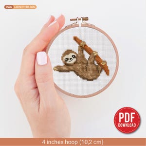 以下が含まれることがあります： 10.2cmの木製刺繍枠に入った、枝にぶら下がる茶色のナマケモノのクロスステッチパターン。デザインは白いアイーダ布に施されています。画像にはWWW.LABPATTERN.COMというテキストと、赤いPDF DOWNLOADバッジが含まれています。